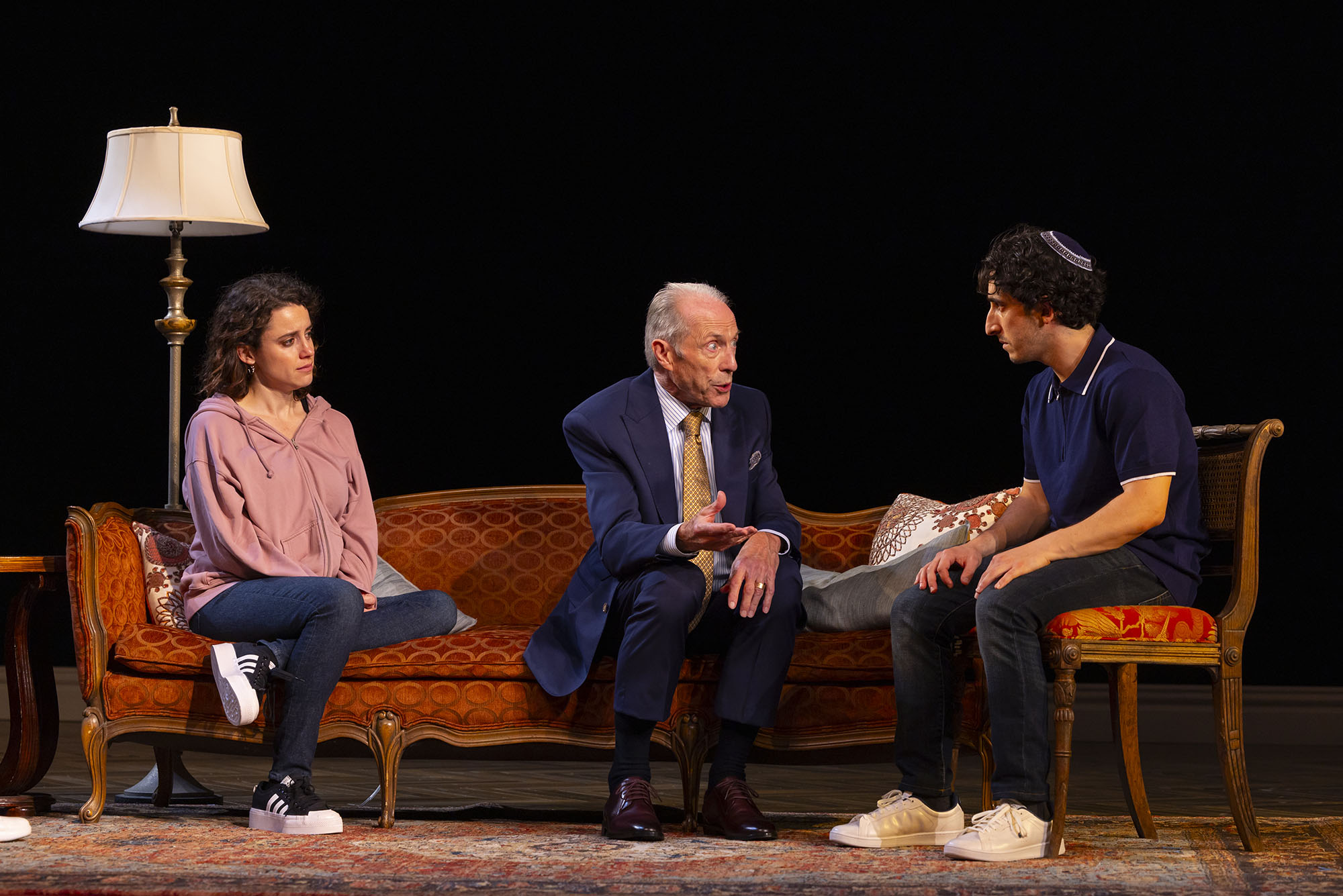 Photo: Three actos sit on a couch, in the middle of a scene. On the far left, a woman with long brown hair sits with one of her legs folded beneath herself. She wears jeans and a pink zip-up hoodie. Next to her, in the middle, is an older man in a navy suit and brown loafers. He is in the middle of talking to the man on the farthest right, a younger gentleman wearing jeans, a navy polo, and a kippah. The couch is orange.
