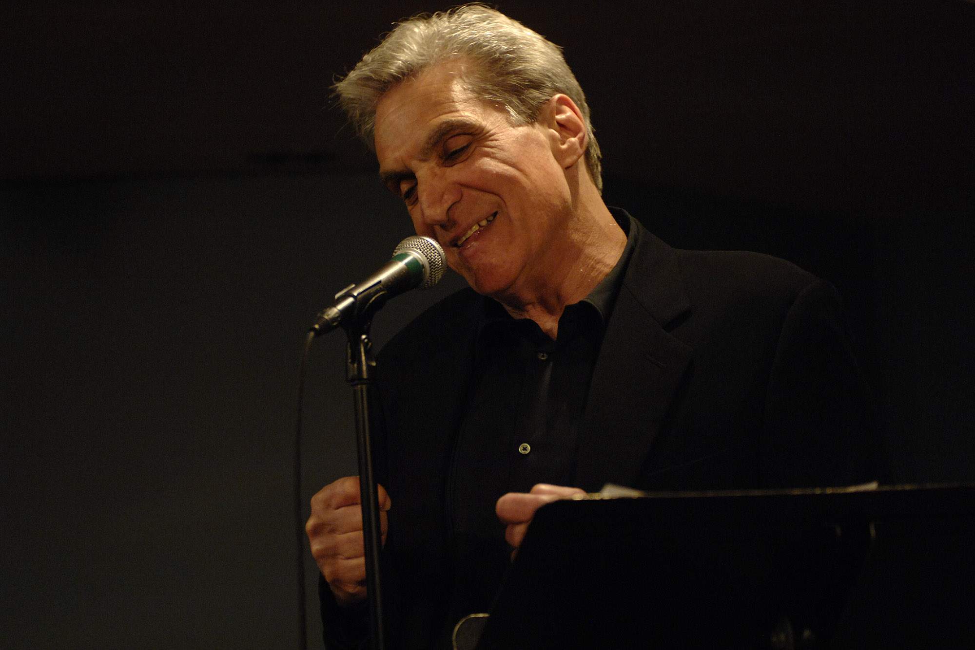 Photo: Poet Robert Pinsky, a white man with a full head of gray hair, stands at a mic with a black button down as he reads his poetry. A single spotlight illuminates him.