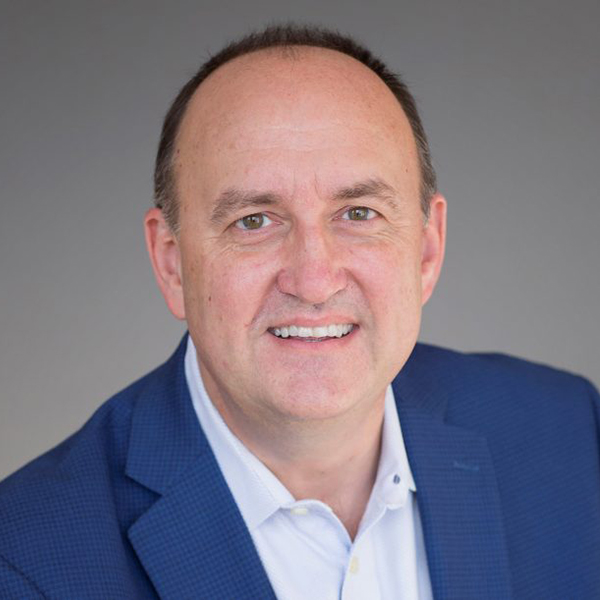 Photo: A white man with a blue blazer and a white button up smiles for a headshot with a plain gray background.