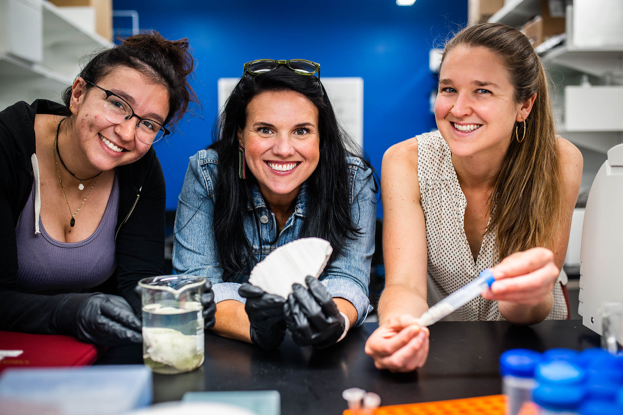 Photo: Group photo of Assistant Professor of Biology, Sarah Davies (center) and PhD student, Maria Valadex Ingersoll (CAS 23) and Dr Hannah Aichelman (right) (CAS 23). The three women pose in a lab as Davies holds up a sample of bleached coral and another sample of algae rests on the lab tables in front of Ingersoll.
