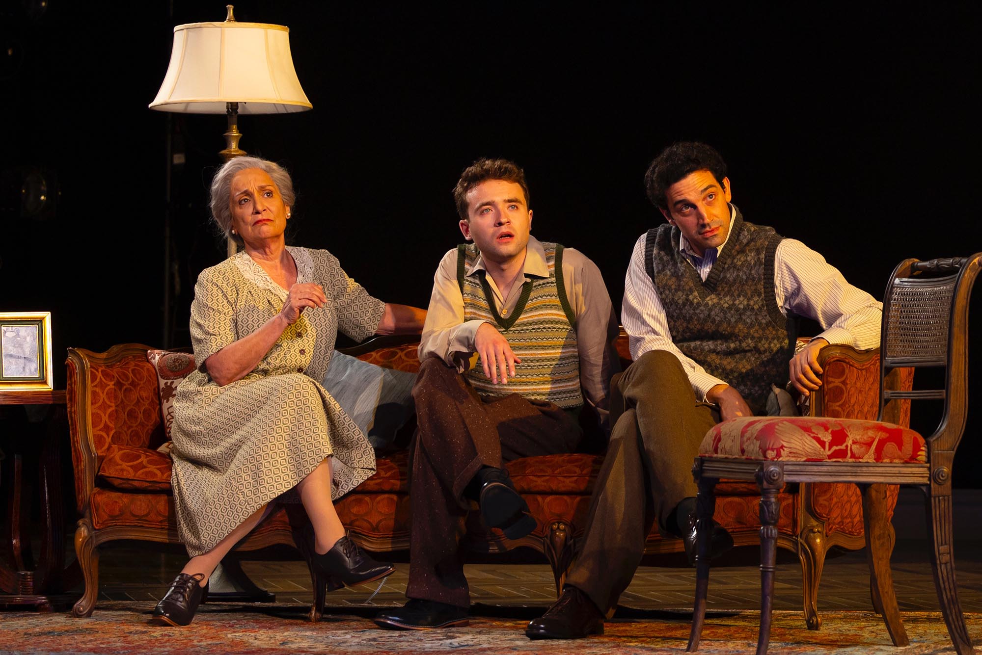 Photo: Three individuals sit on a couch, looking to the far right, off stage. On the far right, an older woman with gray hair and a pattered smock dress. In the middle, a younger man with short brown hair, a stripped vest, brown slacks, and a light button down. And on the farthest right, a man with short brown hair, a patterned vest, brown slacks, and a white button up. Their expressions are intrigued.