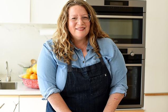 Photo: Adeena Sussman, a white woman with curly brown-blonde hair and wearing a light blue collared shirt and black apron, smiles as she poses for a photo. Her hands work on some pastry dough on the counter in front of her as she smiles in a bright kitchen.