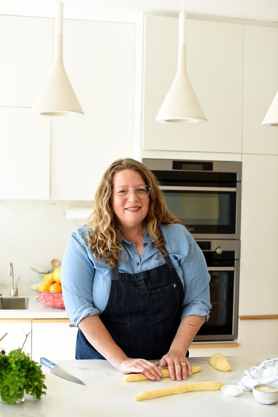 Photo: Adeena Sussman, a white woman with curly brown-blonde hair and wearing a light blue collared shirt and black apron, smiles as she poses for a photo. Her hands work on some pastry dough on the counter in front of her as she smiles in a bright kitchen.