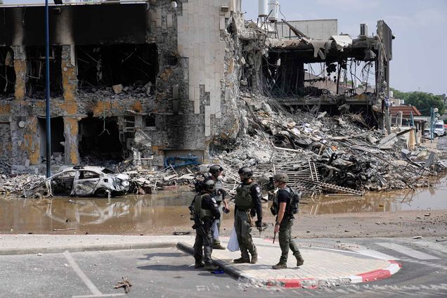 Photo: Israeli police retrieve weapons used by militants outside a police station that was overrun by Hamas gunmen. Four men in army uniforms and holding large rifles talk outside a large building destroyed by a bomb.