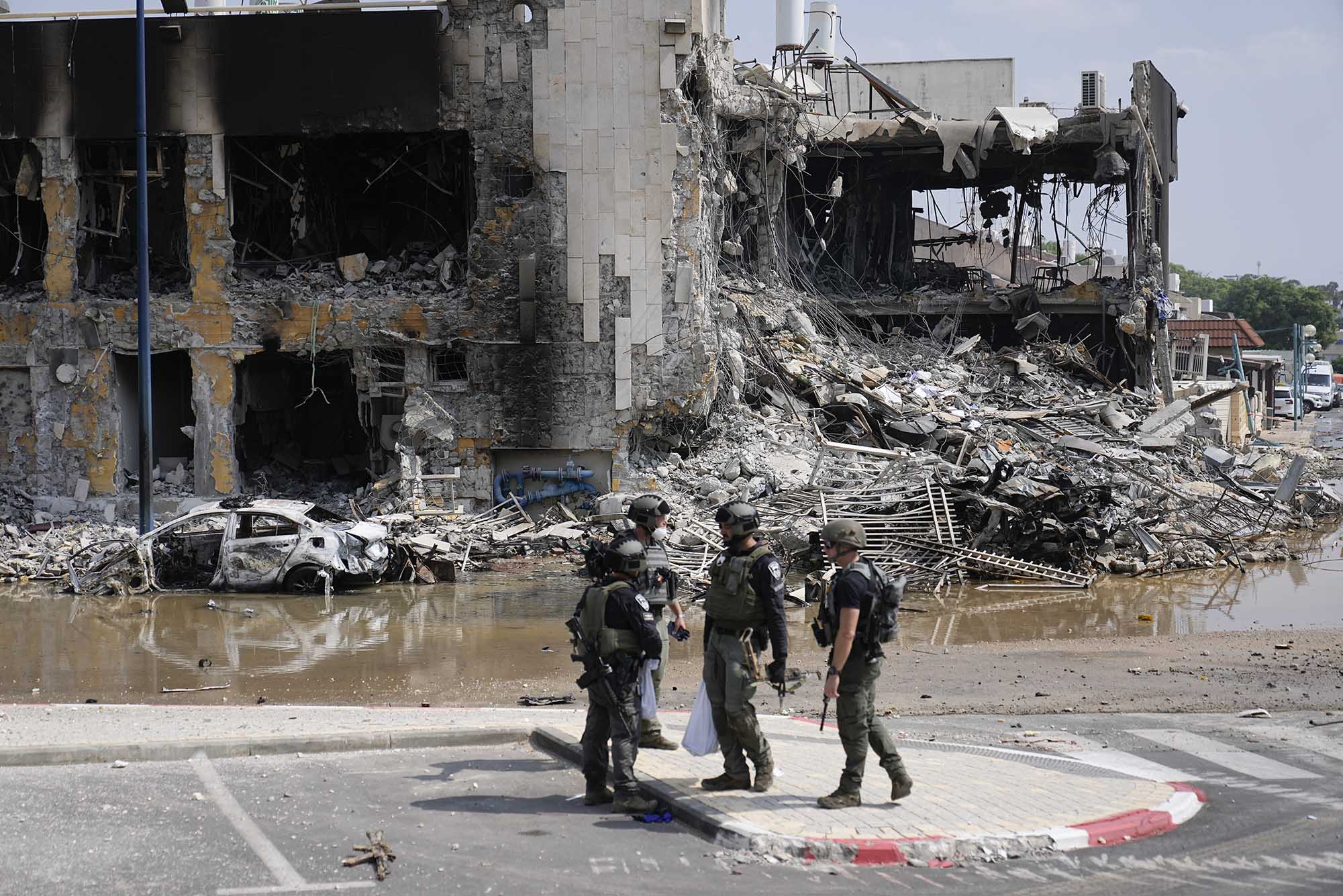 Photo: Israeli police retrieve weapons used by militants outside a police station that was overrun by Hamas gunmen. Four men in army uniforms and holding large rifles talk outside a large building destroyed by a bomb.
