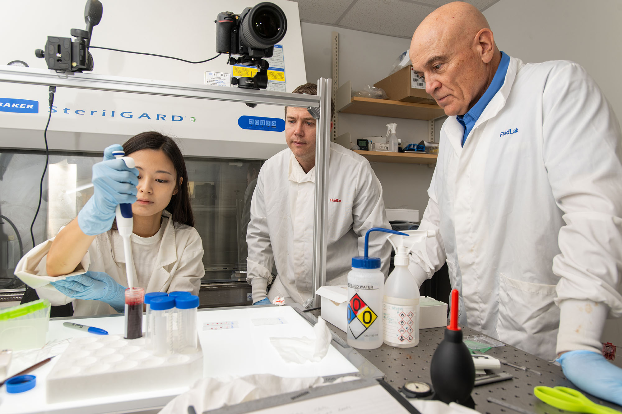 Photo: James Bird (center), a white man with brown hair wearing a white lab coat, Garam Lee (left), an Asian young woman wearing a white shirt and white lab coat, and Kenneth Martin (right), an older white man wearing a white lab coat, work in a lab. Bird and Martin look on as Lee works with blood in the lab.