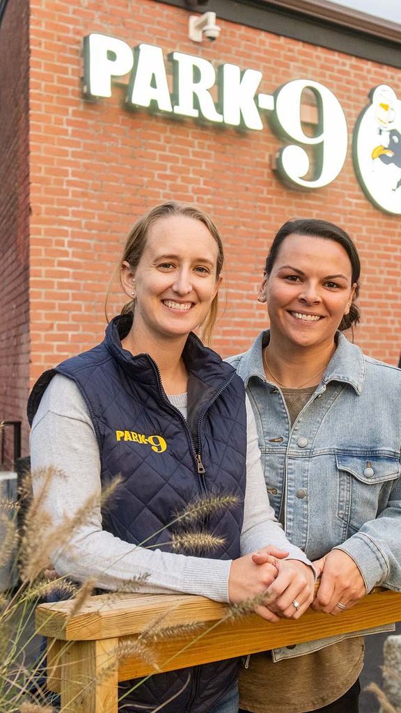 Photo: Emily Gusse, a young white woman wearing a grey long-sleeved shirt and black puffer vest smiles and poses next to Tess Kohanski, a white woman wearing a grey sweater. They both pose in front of a large brick building with a large sign that reads "Park-9".