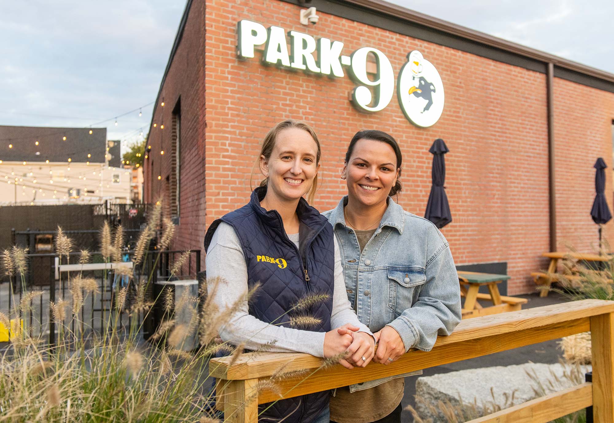 Photo: Emily Gusse, a young white woman wearing a grey long-sleeved shirt and black puffer vest smiles and poses next to Tess Kohanski, a white woman wearing a grey sweater. They both pose in front of a large brick building with a large sign that reads "Park-9".