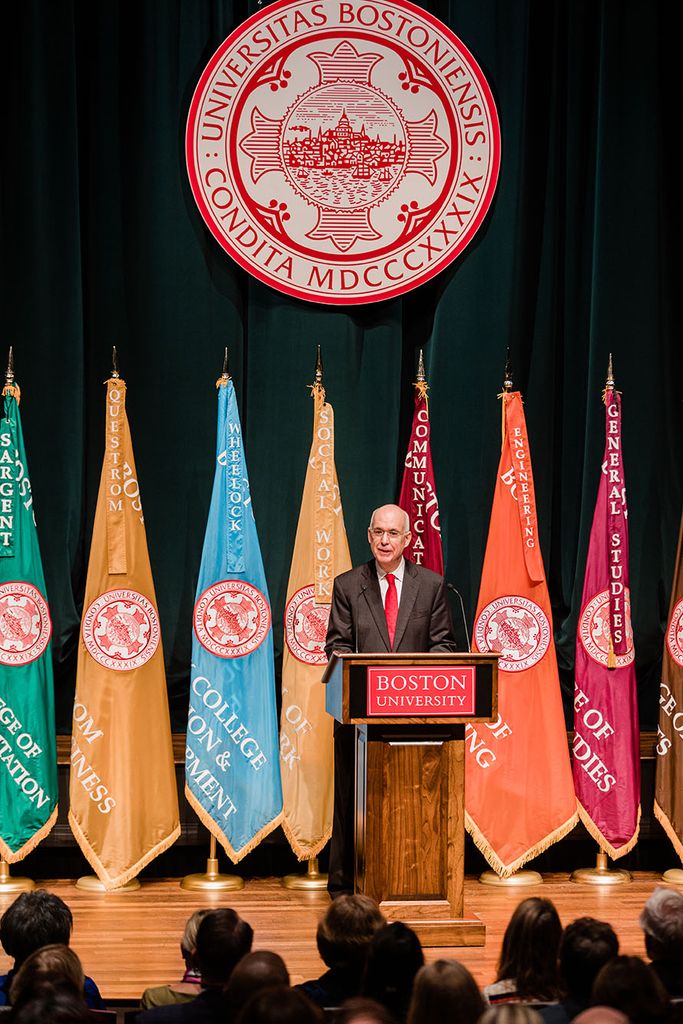Photo: Kenneth Freeman, a White man wearing glasses and a grey suit, speaks into a microphone on a wooden podium. Sign on podium reads "Boston University". Behind him, a row of colorful flags are placed as well as a large BU insignia.