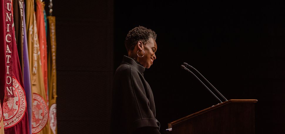 Photo: Dr. Melissa L.Gilliam, a Black woman with a short afro and wearing a black turtleneck sweater, speaks into a microphone on a wooden podium. Sign on podium reads "Boston University". Behind her, a row of colorful flags are placed.