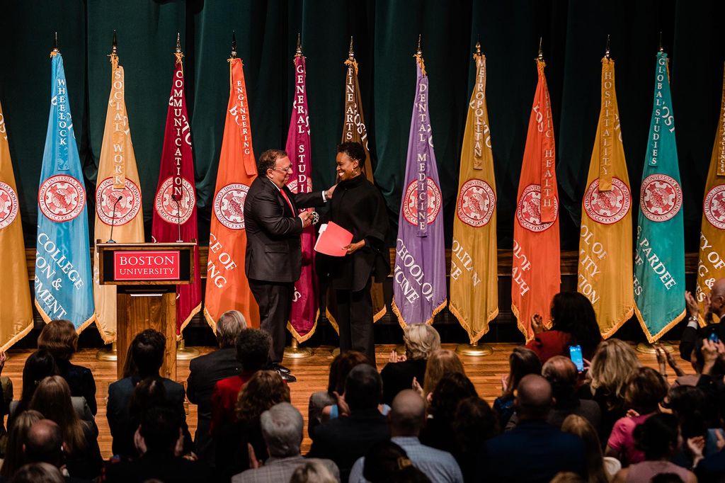 Photo: Ahmass Fakahany, a tan man wearing a black suit, and, Dr. Melissa L.Gilliam, a Black woman with a short afro and wearing a black turtleneck sweater, smile and shake hands on a large stage next to a wooden podium. Sign on podium reads "Boston University". Behind them, a row of colorful flags are placed.