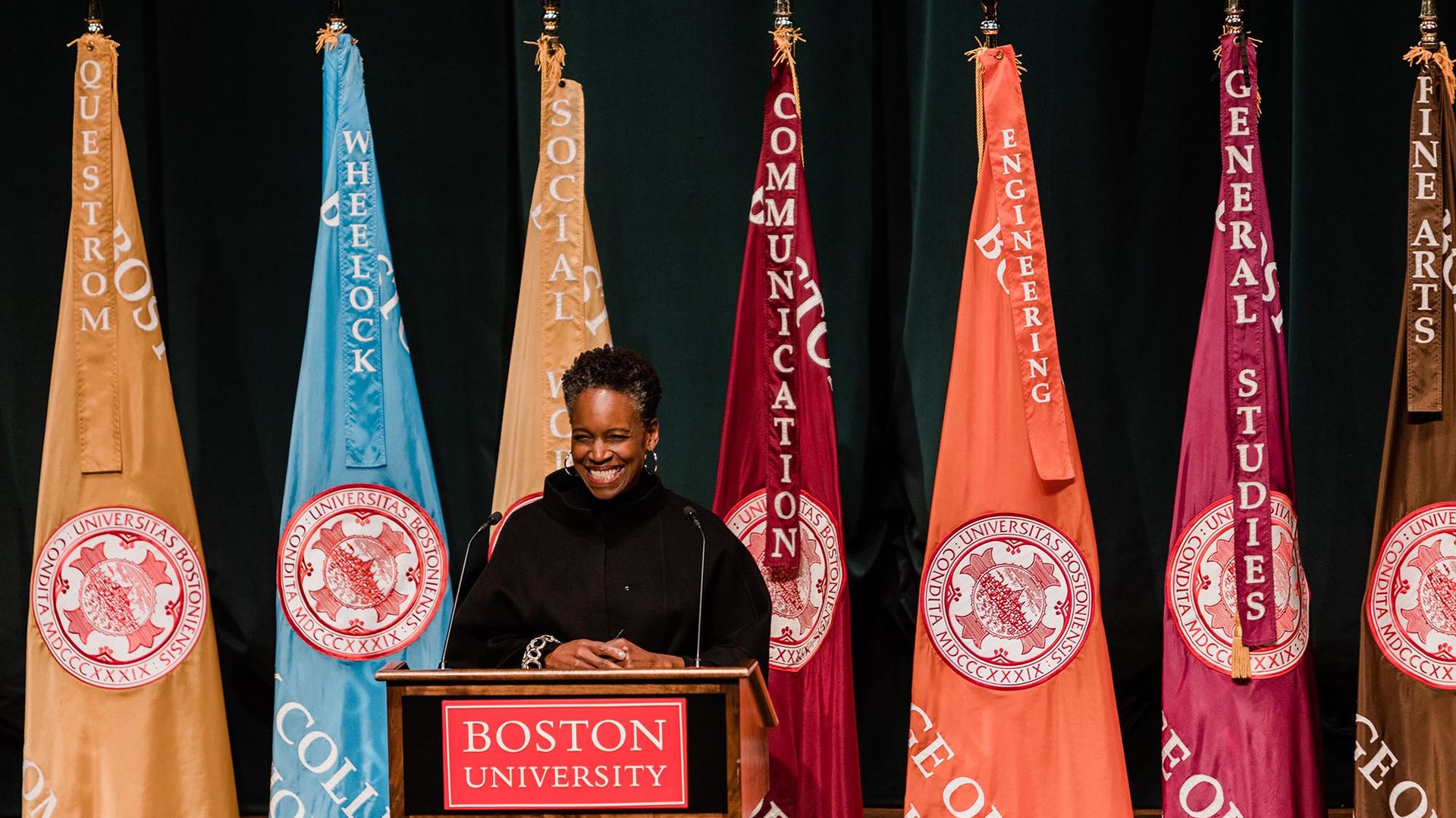 Photo: Dr. Melissa L.Gilliam, a Black woman with a short afro and wearing a black turtleneck sweater, speaks into a microphone on a wooden podium. Sign on podium reads "Boston University". Behind her, a row of colorful flags are placed.