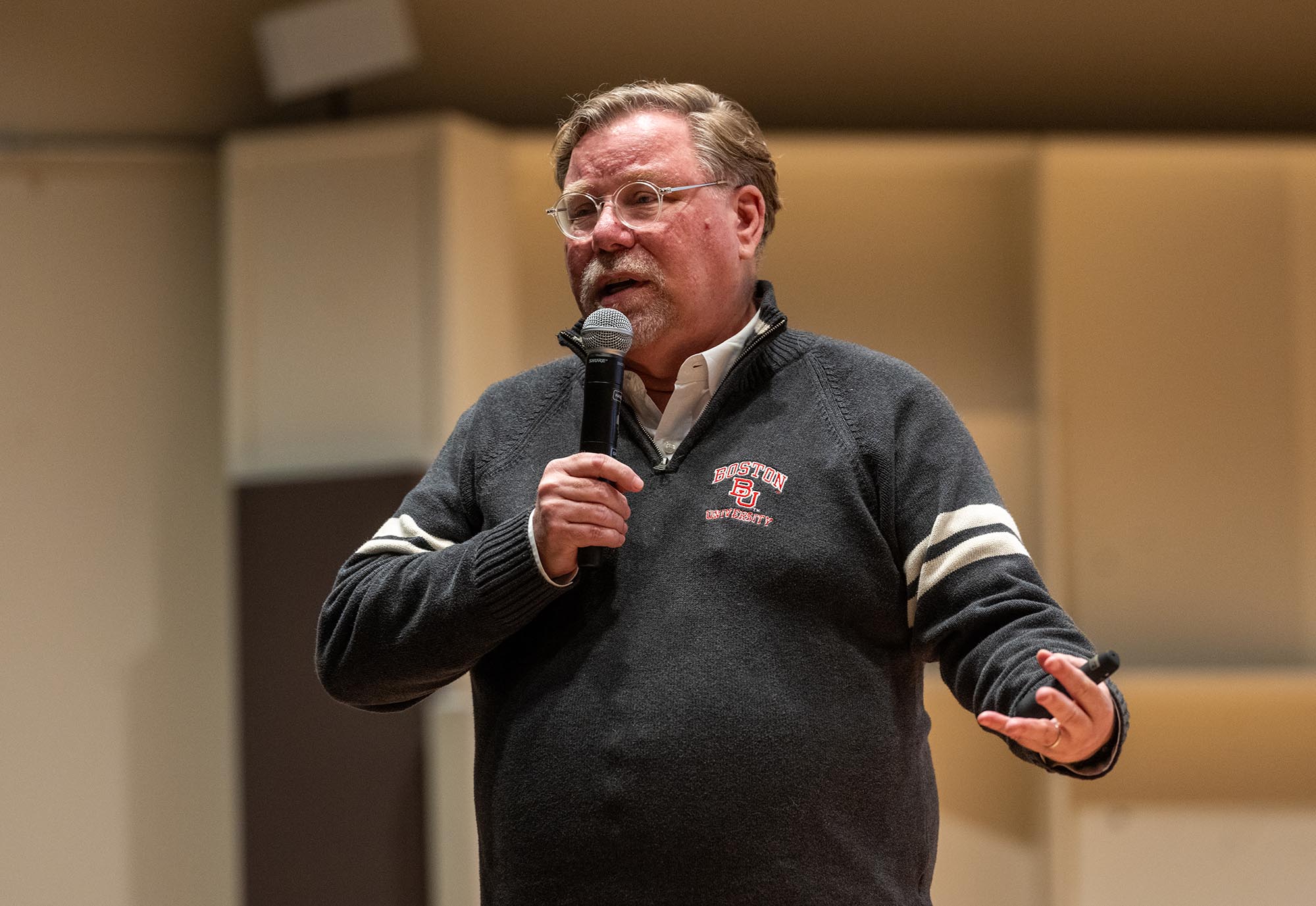 Photo: Gregory Melchor-Barz, a white man with brown-gray hair and wearing glasses and a dark grey long-sleeved shirt, gesticulates as he speaks into a microphone in his hand.
