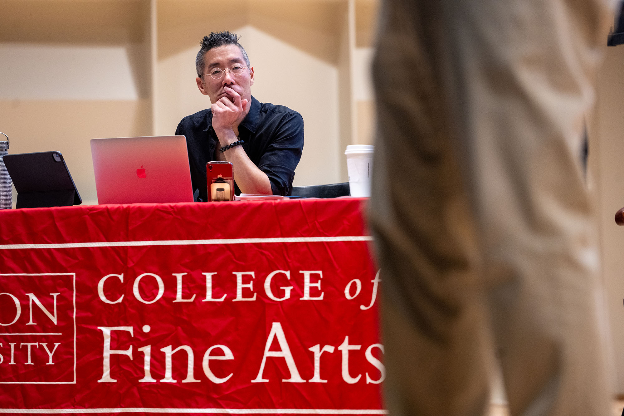 Photo: Kinh Vu, an Asian man with peppered black hair and wearing a black turtleneck, sits at a table with a large red banner on the front that reads "College of Fine Arts".