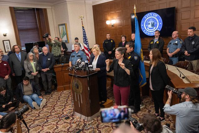 Photo: wide shot of Maine Governor Janet Mills speaking during a press conference about the mass shooting on October 26, 2023 in Lewiston, Maine. A white woman in business attire stands at a wooden podium front and center of the room. Behind her, police officers and officials stand. To her right, an ASL interpreter signs. In front of her, various journalists and photographers write, take photos, and listen.