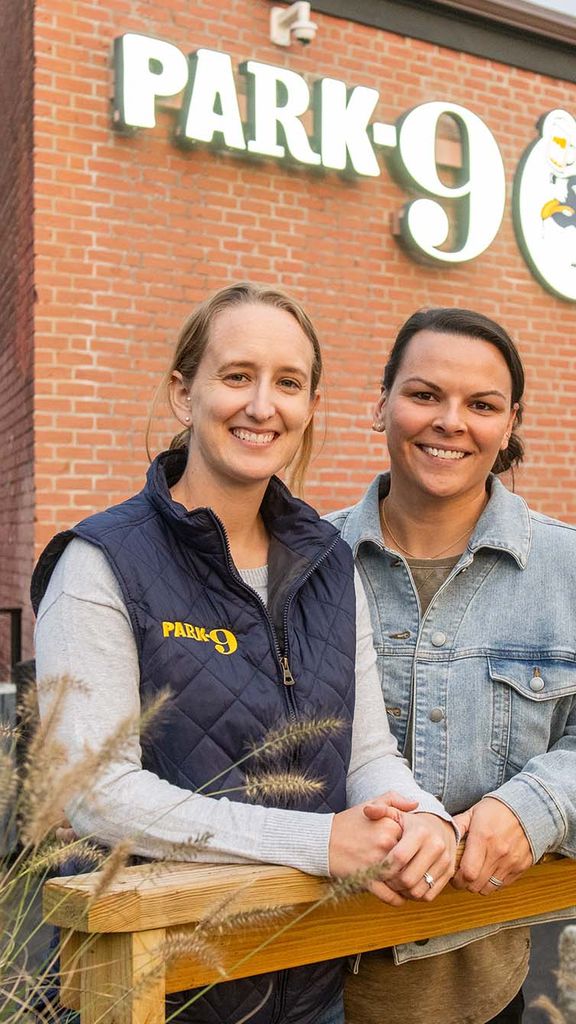 Photo: Emily Gusse, a young white woman wearing a grey long-sleeved shirt and black puffer vest smiles and poses next to Tess Kohanski, a white woman wearing a grey sweater. They both pose in front of a large brick building with a large sign that reads "Park-9".