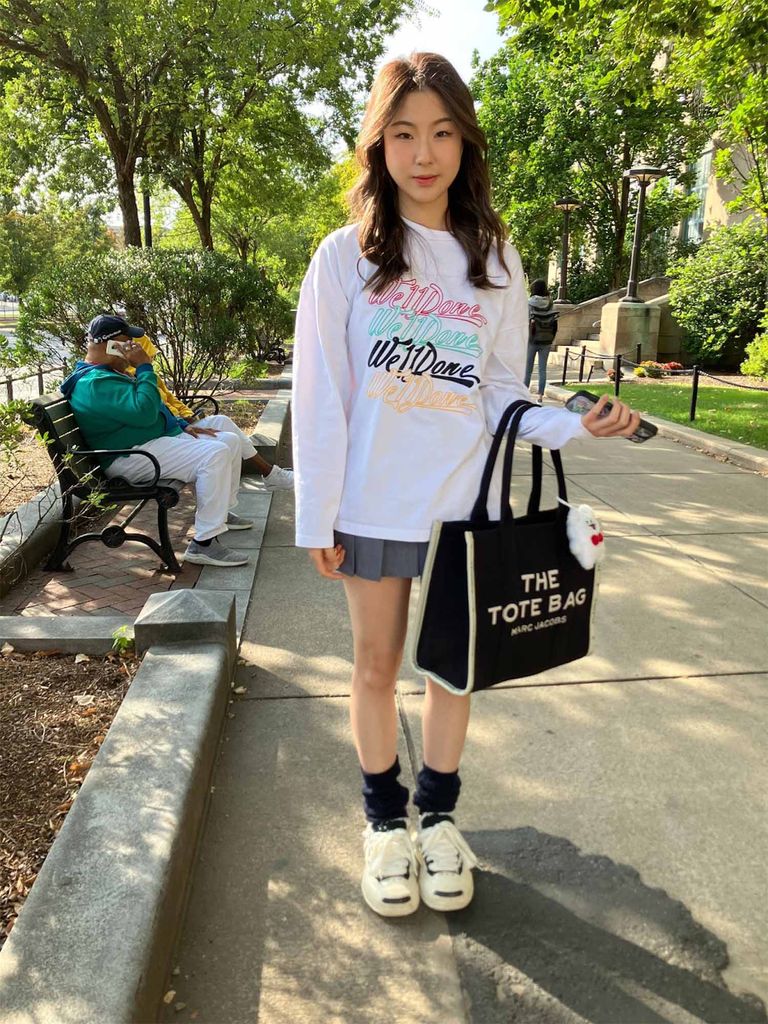 Photo: A student wearing a skirt, white shoes, and a sweatshirt carries a bag that says "The Tote Bag" as they stand on a city sidewalk