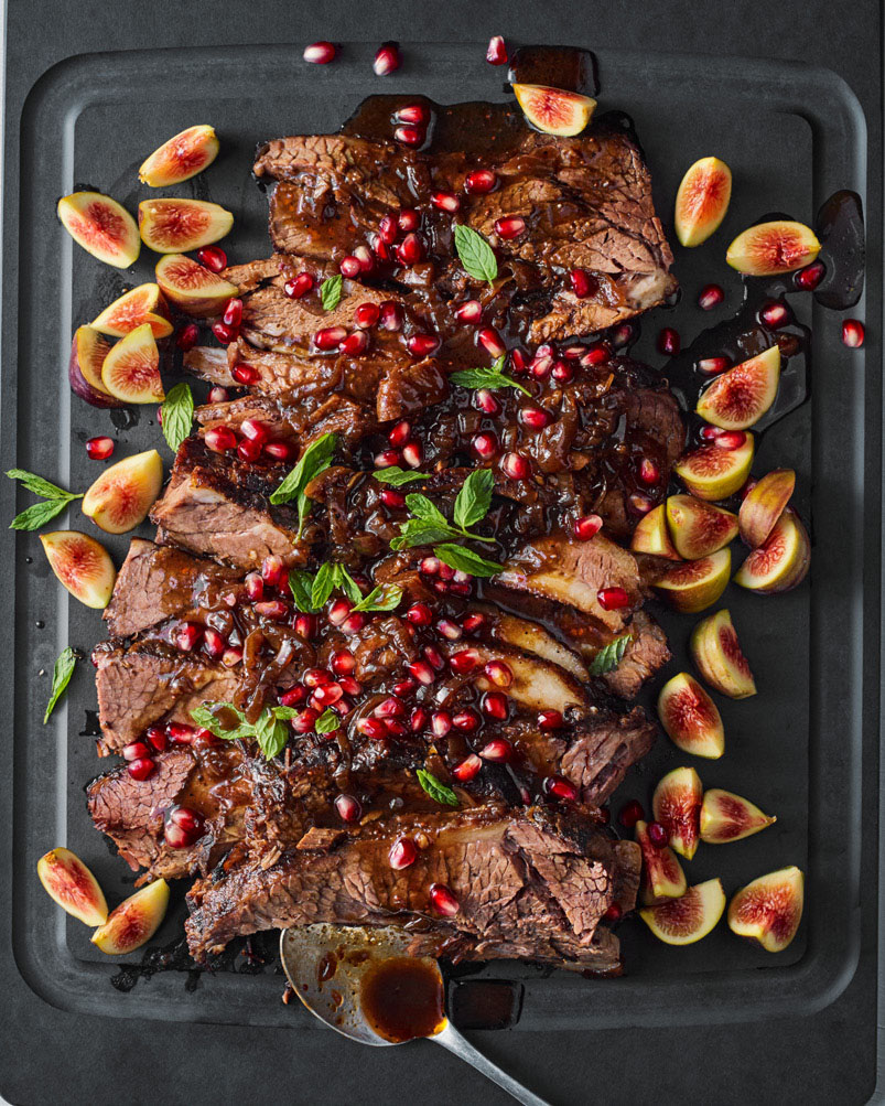 Photo: Overhead shot of a nicely plated fig and pomegranate brisket. Brisket and fig slices are artfully arranged on a large black platter. The dish is topped with a brown sauce, pomegranate seeds, and green mint leaves.