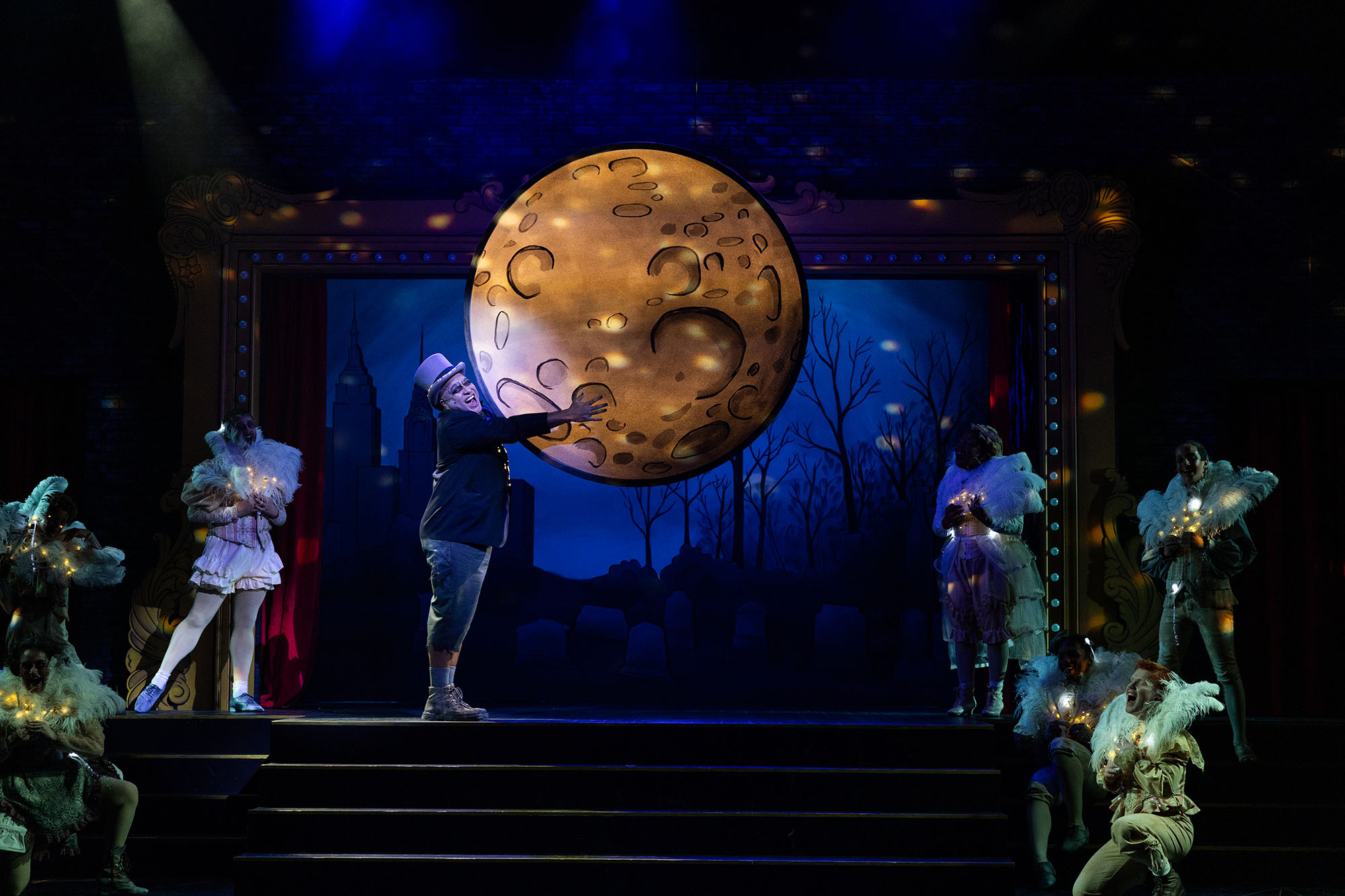 Photo: An man with Ashy makeup and wearing a large blazer, jeans, and a grey top hat, smiles and sings excitedly as he grabs onto a large moon hanging down on set. Dancers wearing large white feathers and white outfits sing and smile on the sidelines of the stage.