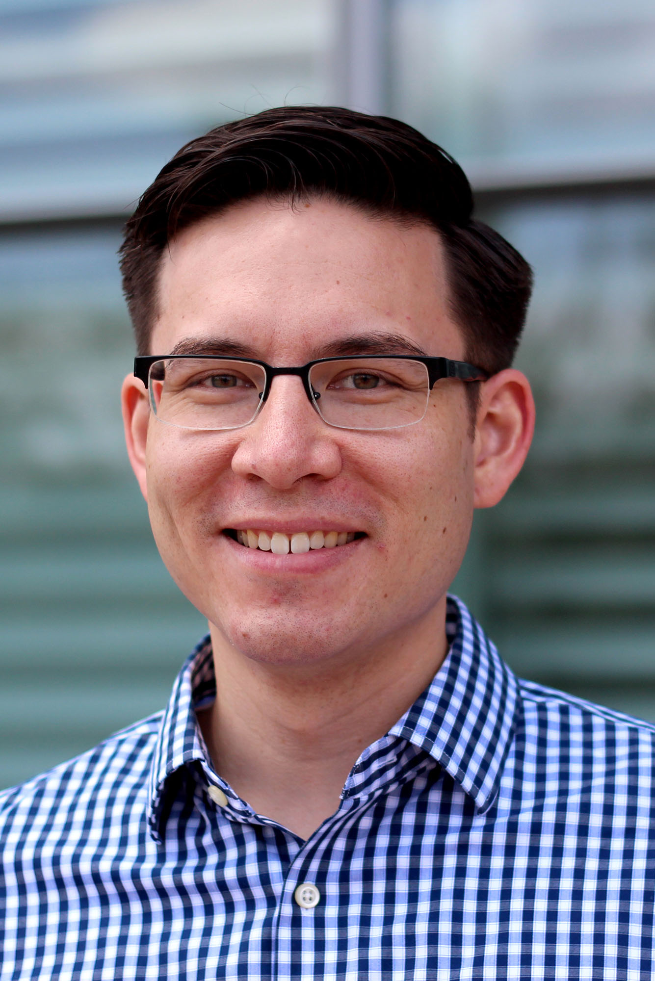 Photo: A headshot of Alex Green, a man with short dark hair, rectangular black glasses, and a checkered button up. He smiles slightly for the camera.