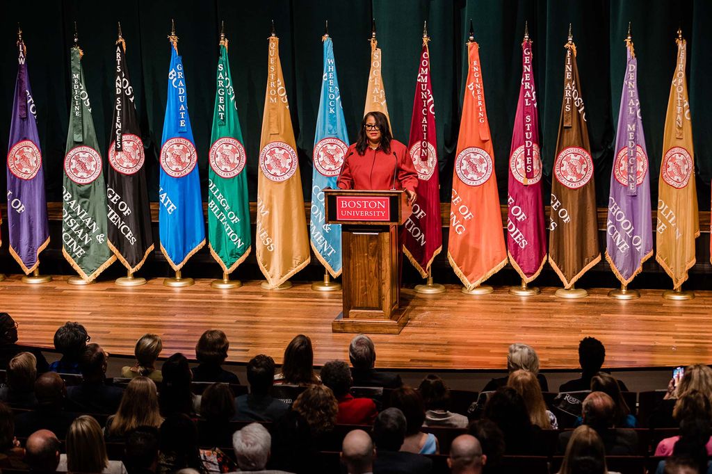 Photo: Antoinette “Tonie” Leatherberry, a Black woman wearing glasses and a long-sleeved red collared shirt, speaks into a microphone on a wooden podium. Sign on podium reads "Boston University". Behind her, a row of colorful flags are placed.