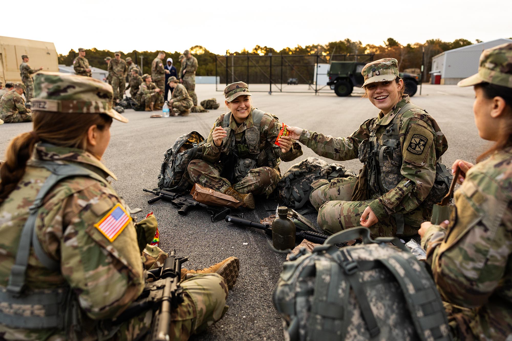 Photo: ROTC Army Cadet, Audrey Soukup (Sargent’25) (second from right) and Dharma Cooper (Pardee’25) share Reeces Pieces candy while they wait with their platoon. A small group of young women dressed in army ROTC gear sit on the ground and share candy with each other.