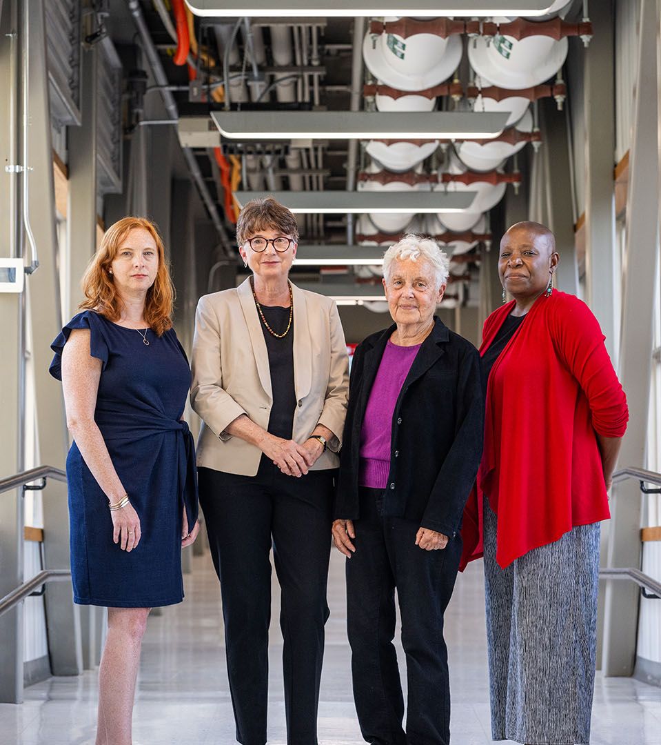 Photo: (from left) Kimberly Bertrand, a white woman with auburn hair and wearing a navy dress, Julie R. Palmer, a white woman wearing glasses, black blouse and pants, and tan blazer, Lynn Rosenberg, an older white woman with white hair and wearing a purple blouse, black blazer, and black pants, and Yvette Cozier, a bald Black woman wearing a black blouse, red cardigan, and grey skirt. All four stand and pose for a photo in a wide, brightly lit hallway.