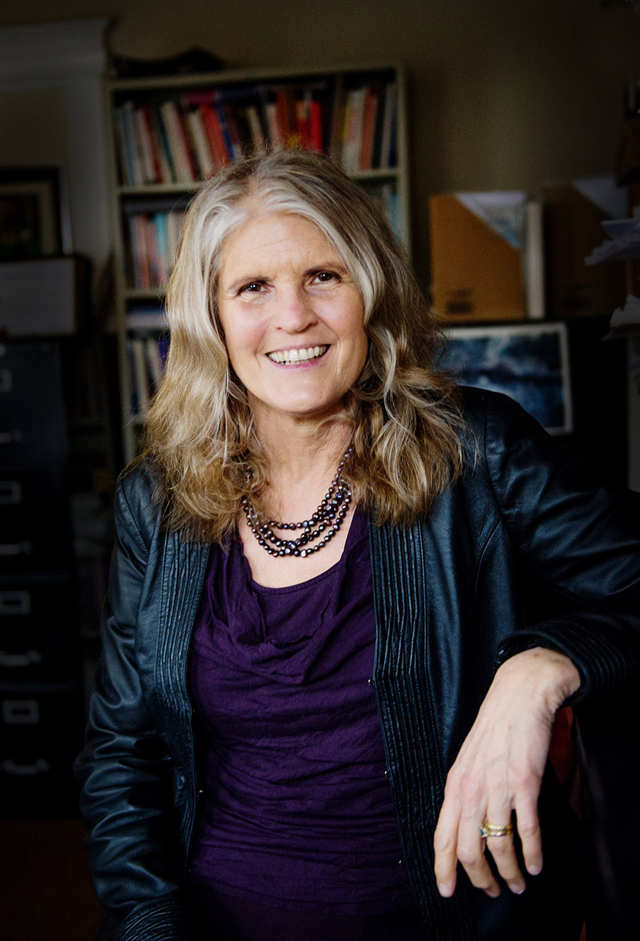 Photo: Cathie Jo Martin, a white woman with curly blonde hair and wearing a dark purple blouse and dark blue jacket smiles and poses with one arm resting on the armrest of the chair she sits in. A bookcase filled with books can be seen in the background.