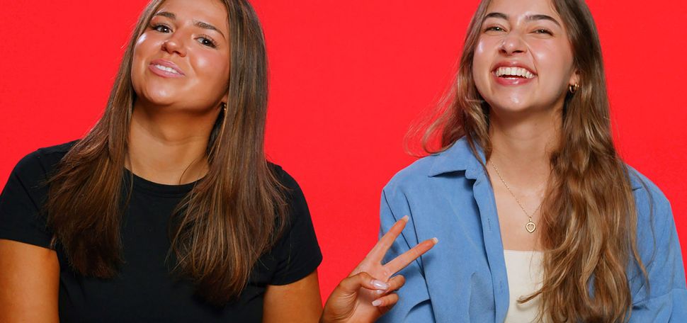 Photo: Two white young women in casual wear smile and pose for the camera in front of a bright red background. They sit at a small, round wooden table with a small pile of red cards on it.