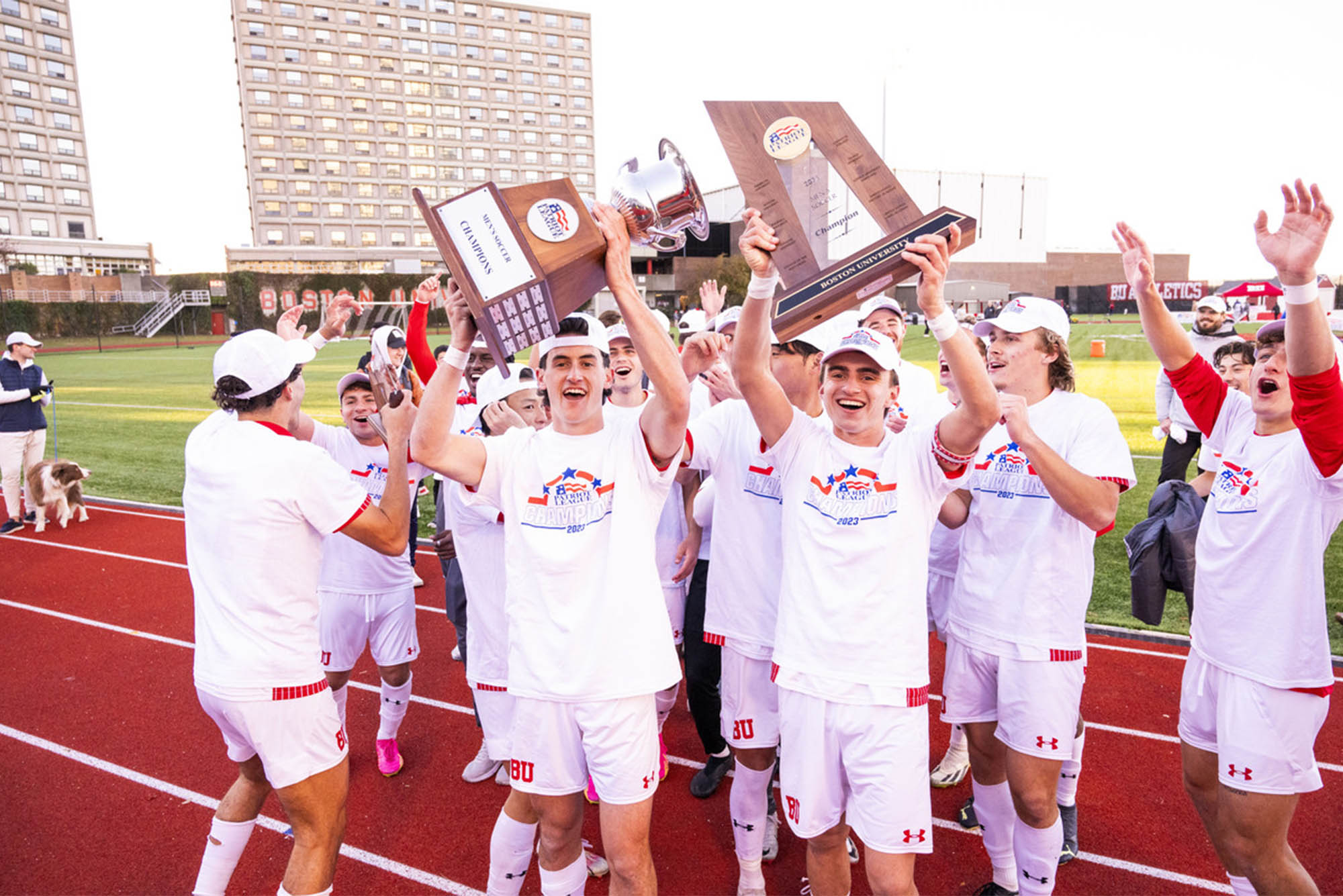 Photo: BU Men's soccer players raise the Patriot League trophy after a large win. Players are wearing white jerseys and standing on a soccer field