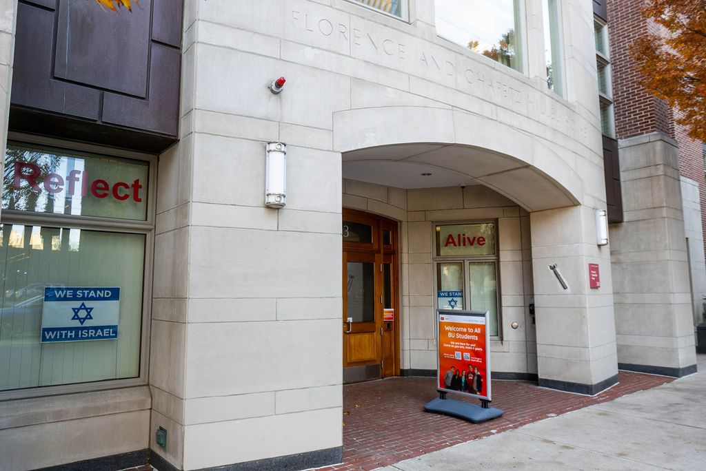 Photo: Exterior shot of BU's Hillel House. A grey brick building with wooden doors is shown. A red sign saying "Welcome to all BU Students" is placed in front of the door near the sidewalk and an Israeli flag can be seen in the left window of the building.