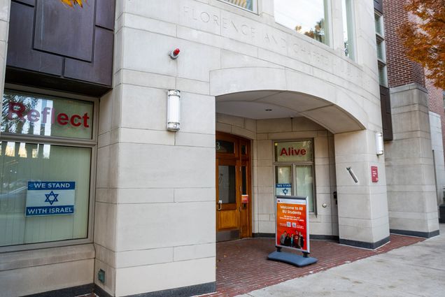 Photo: Exterior shot of BU's Hillel House. A grey brick building with wooden doors is shown. A red sign saying "Welcome to all BU Students" is placed in front of the door near the sidewalk and an Israeli flag can be seen in the left window of the building.