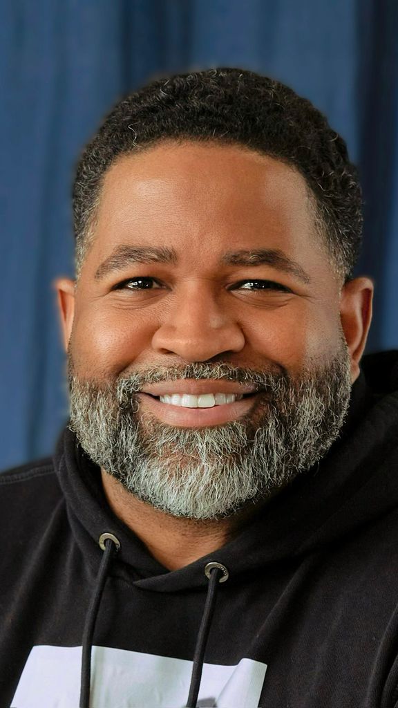 Photo: Jamil Smith, a man with a beard, smiles for the camera. He is wearing a black hoodie and is in front of a blue curtain background.