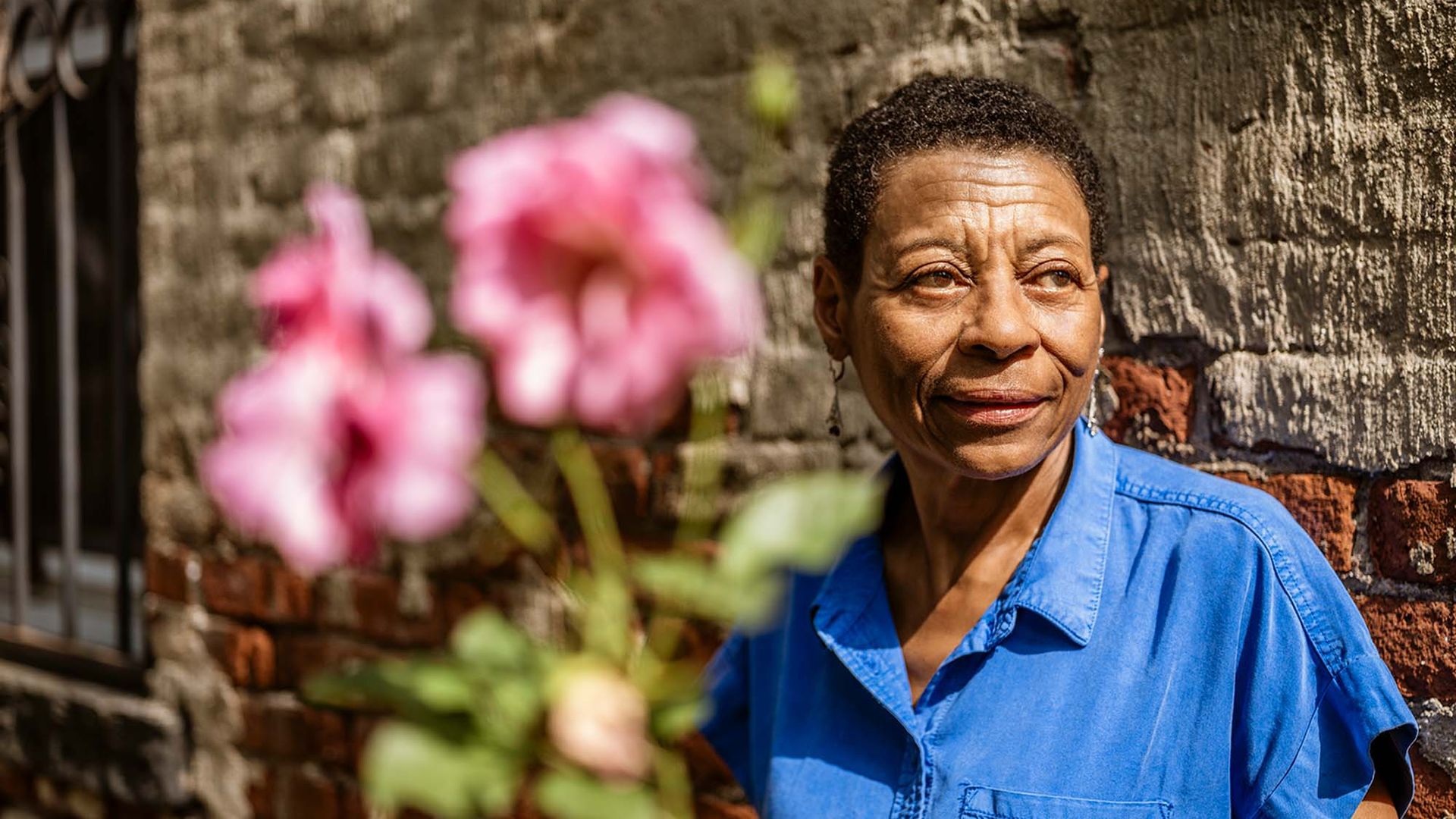 Photo: Kim Bressant Kibwe, an older Black woman with short, black hair and wearing a blue, button-up collared shirt smiles and looks to the right as she poses in front of a brick wall. A blurred pink flower is shown in the foreground.