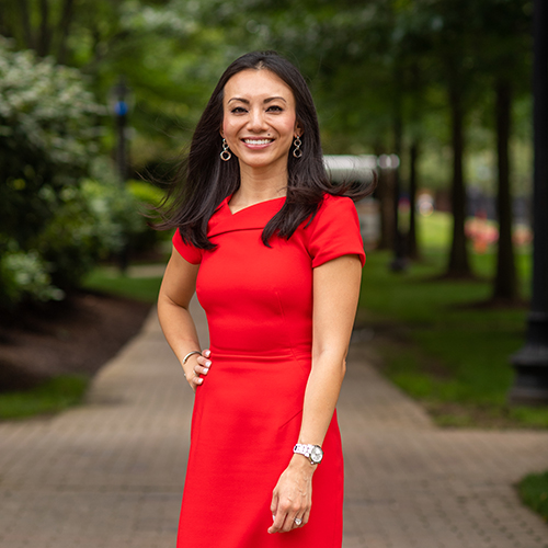 Photo: Monica Wang, an Asian woman with long black hair and wearing a short sleeve red dress, smiles and poses with one arm on her hip. She stands outside on a park walkway, as blurred greenery can be seen behind her.