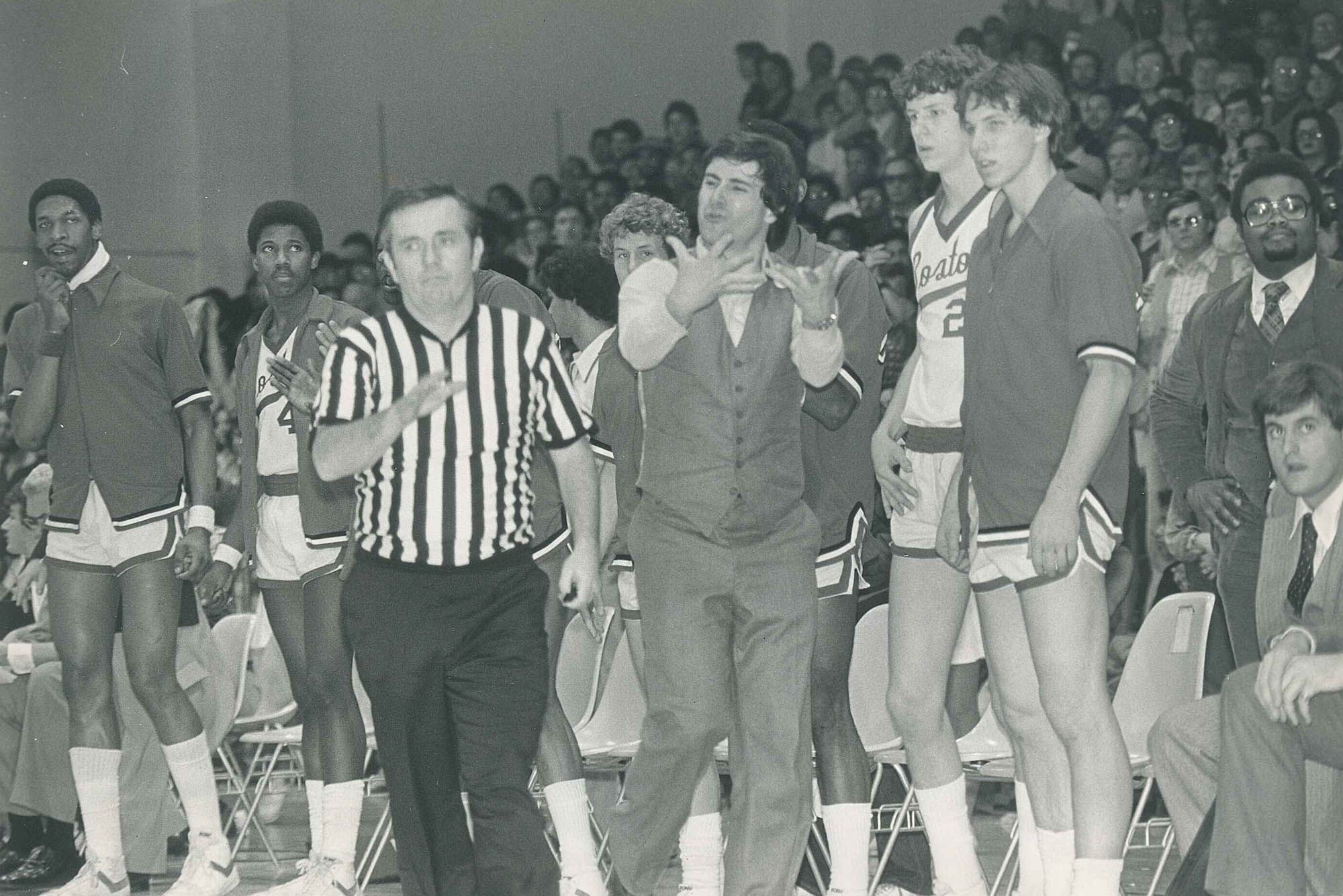 Photo: A black and white photo of Rick Pitino coaching from the sidelines of a BU mens basketball game. He and the coaches stand as they watch the not shown game. Tot he left, a referee is shown running down the court.