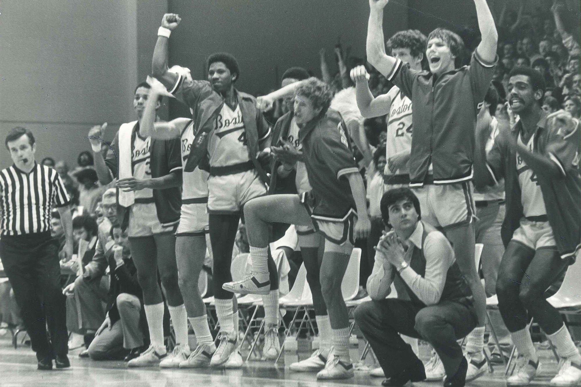 Photo: A black and white photo of Rick Pitino coaching from the sidelines of a BU mens basketball game. He and the athletes react excitedly to a play on the court as they watch the not shown game. Tot he left, a referee is shown running down the court.