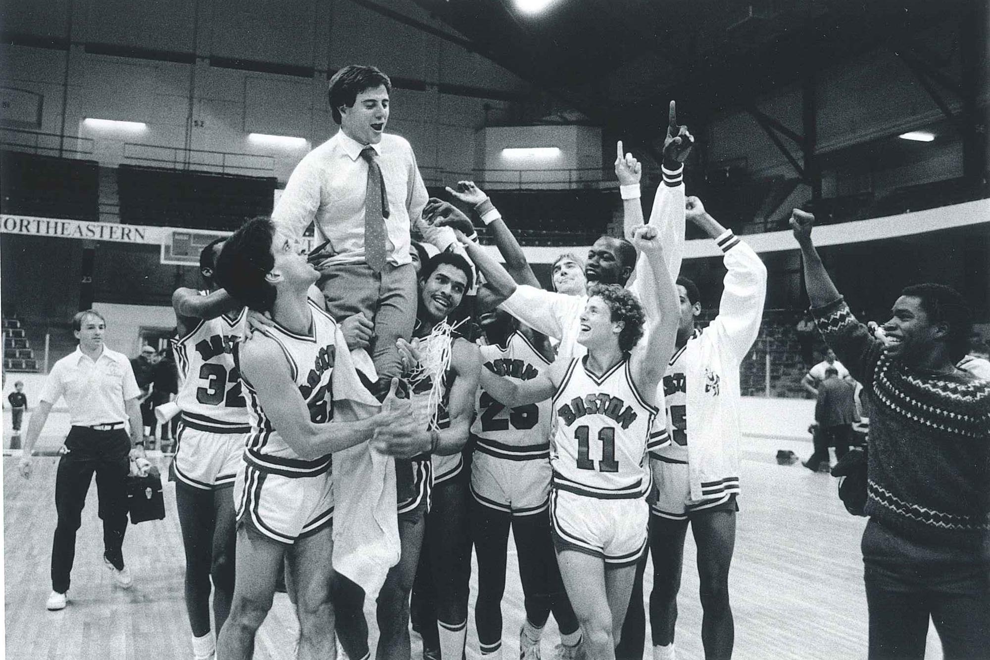 Photo: A black and white photo of Rick Pitino celebrating with the BU men's basketball time he coached. Members of the team hoist him on their shoulders as they group up and cheer on the basketball court.