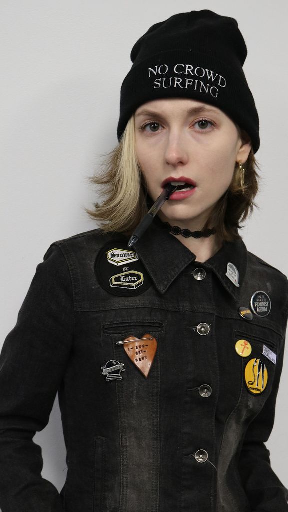 Photo: A woman with short blonde hair wearing a black beanie that says "No Crowd Surfing" and a jacket covered in pins