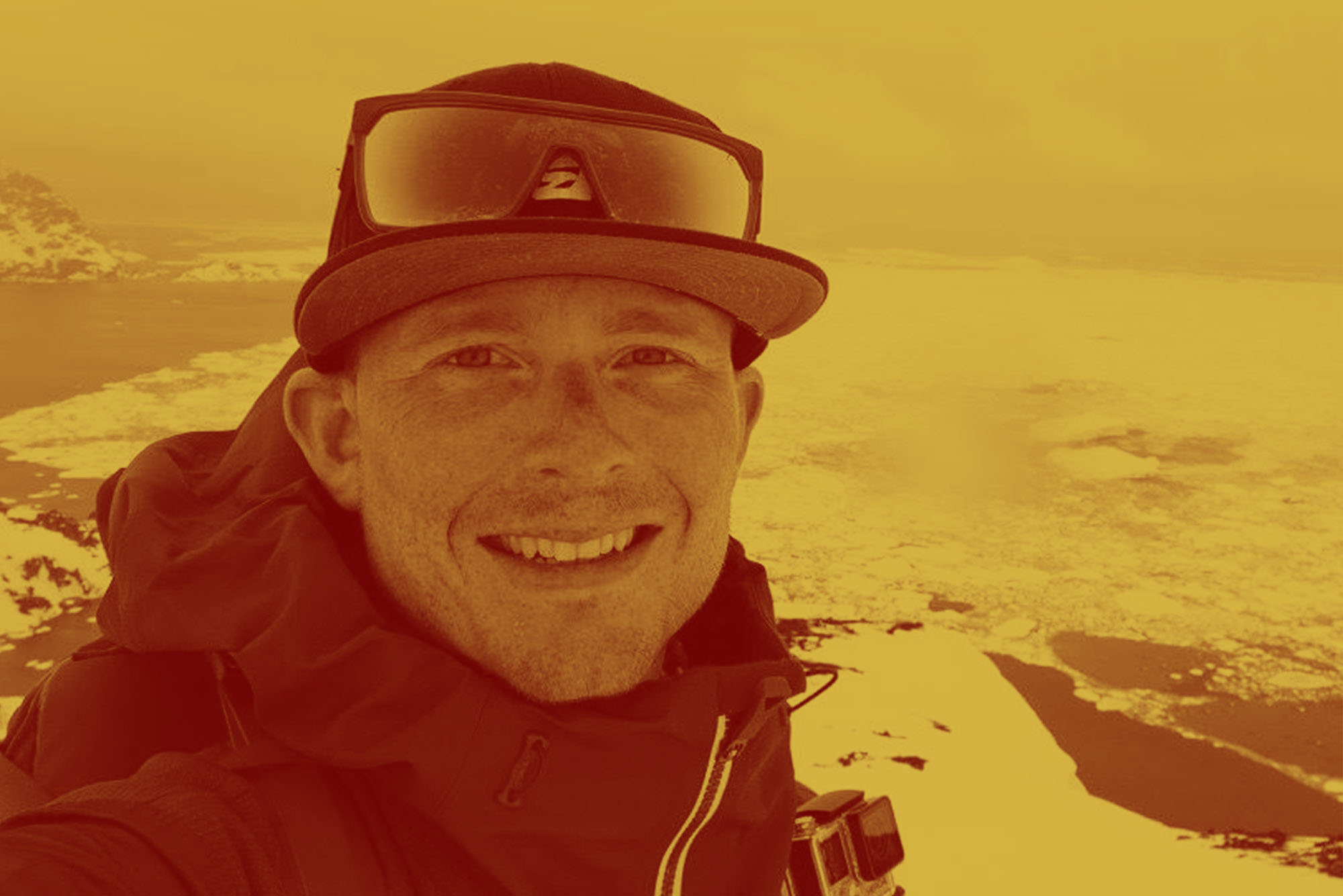 Photo: A White man wearing a hat and sunglasses stands atop a snowy mountain. The photo has an orange overlay.