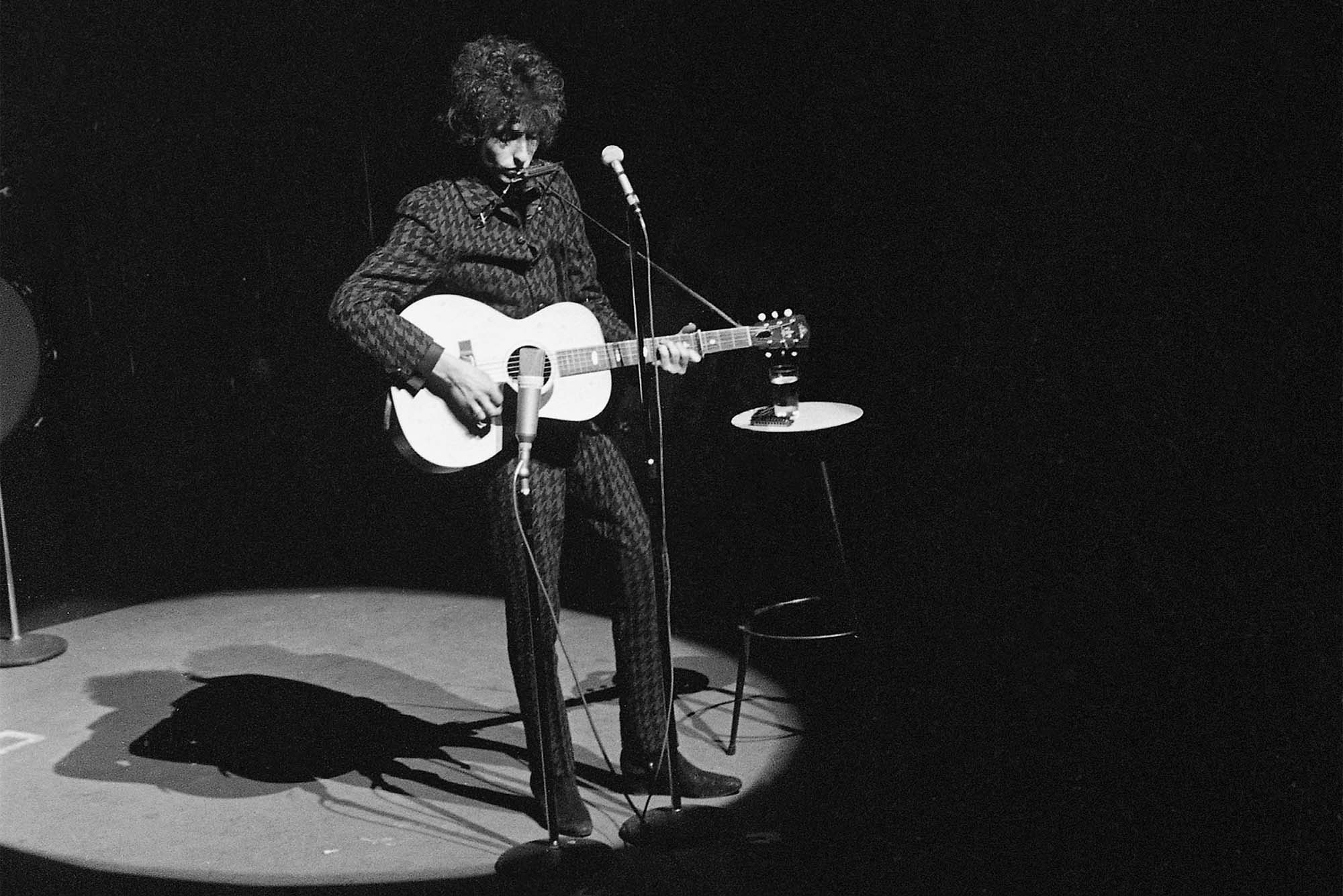 Photo: A black and white photo of a young Bob Dylan in Paris in 1966, on his first tour since “going electric” at the Newport Folk Festival. A young man with curly hair stands on stage under a bright spotlight and play the guitar. He stands in front of a standing microphone.