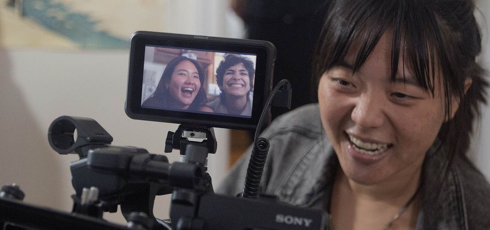 Photo: A young Asian woman is shown filming using a large black film camera. The small display on the camera shows two young adults in the midst of being filmed for a scene.