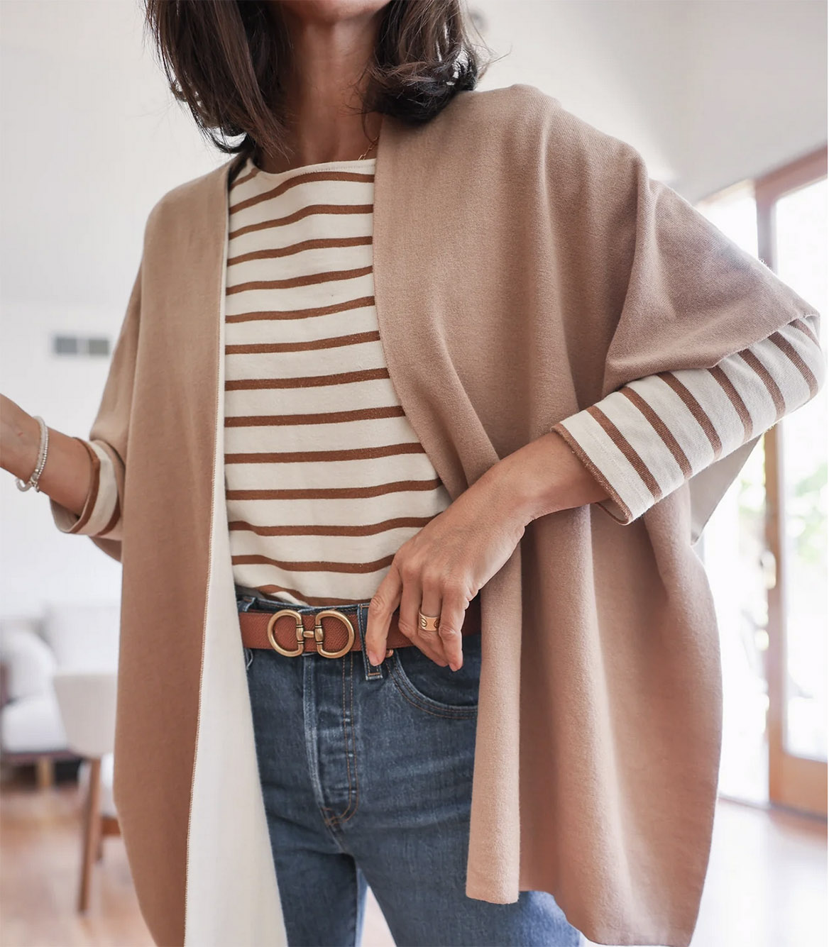 Photo: Zoomed in photo shows a person wearing a white and brown striped shirt, beige reversible cardigan, and jeans. They pose in a brightly lit space.