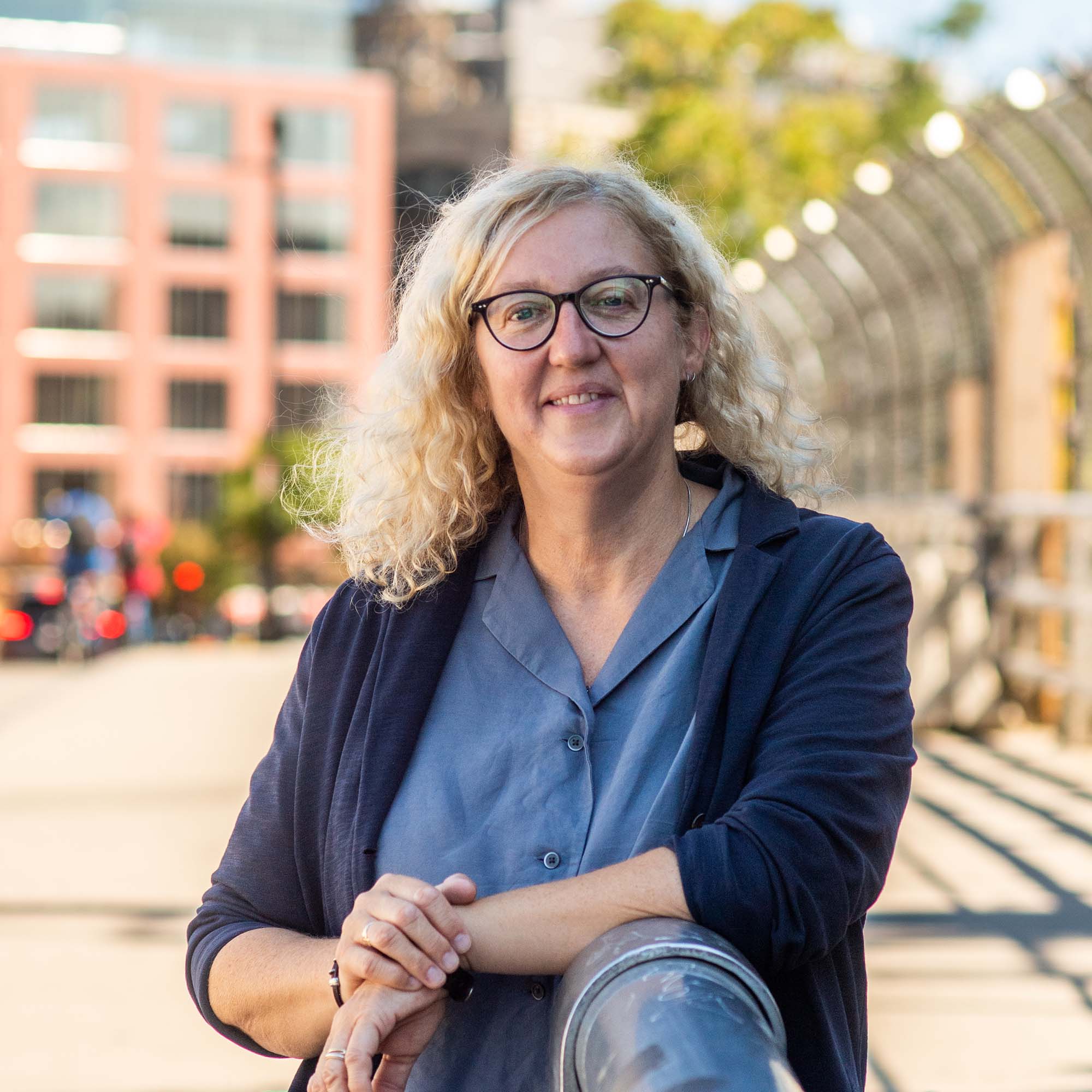 Photo: A white woman wearing a blue shirt and black cardigan stands outside on a sunny day with a smile on their face