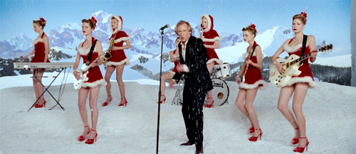 Movie gif. This was from the movie Love Actually and actor Bill Nighy is singing and spinning around in front of a microphone. Behind him is a winter themed set with fake snow and mountains. There are backup dancers wearing santa dresses, dancing in sync.