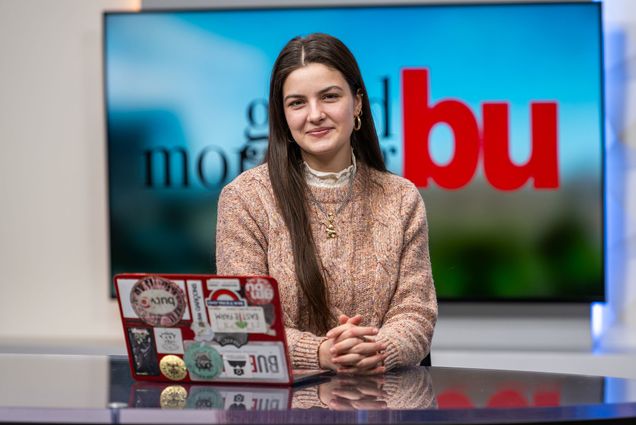 Photo: Alex Dowd, a woman, smiles at a news desk with a red laptop with a TV screen that reads "GOOD MORNING, BU" behind them.