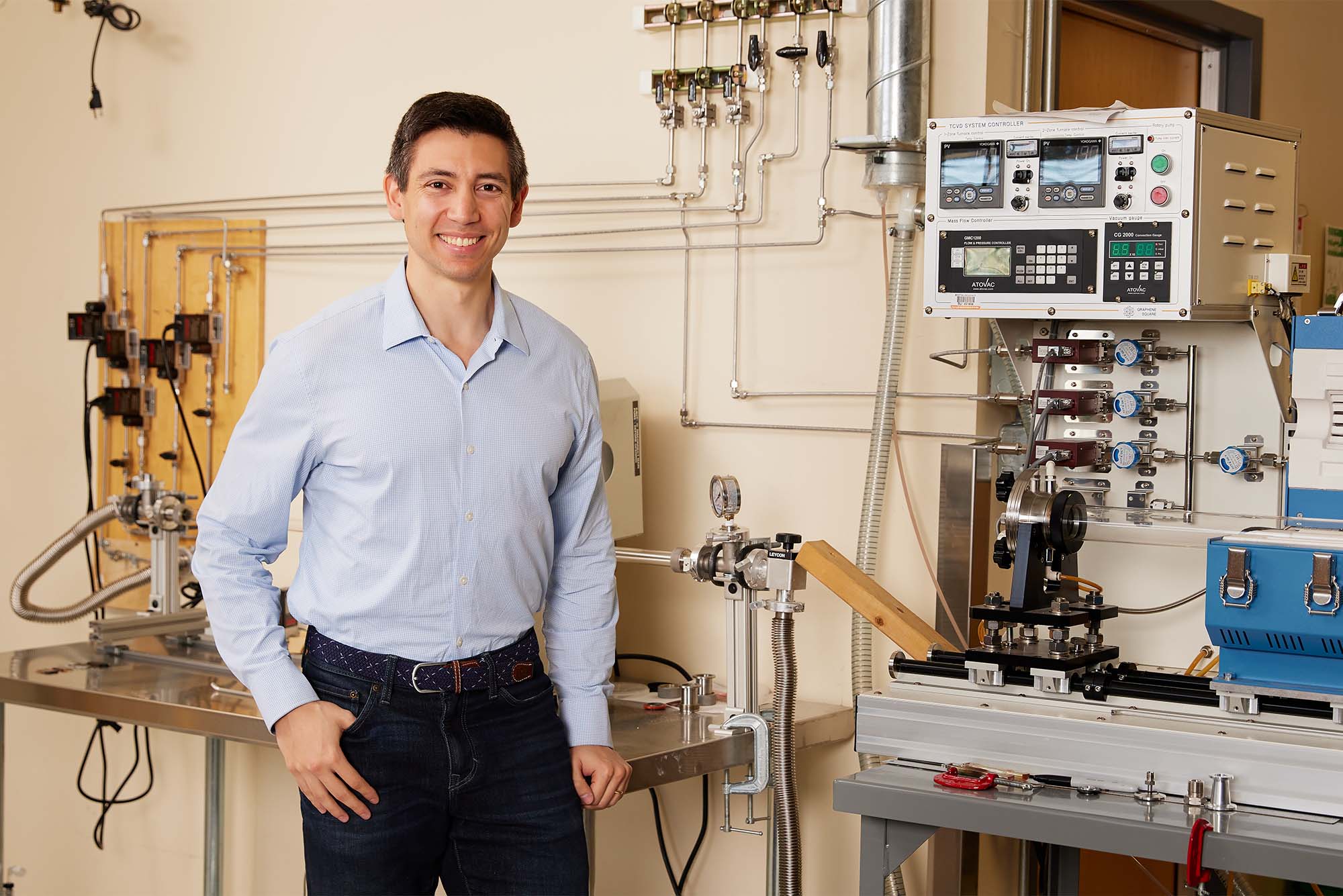 Photo: A man in a collared shirt with an elegant belt stands in front of complex machinery in a lab environment