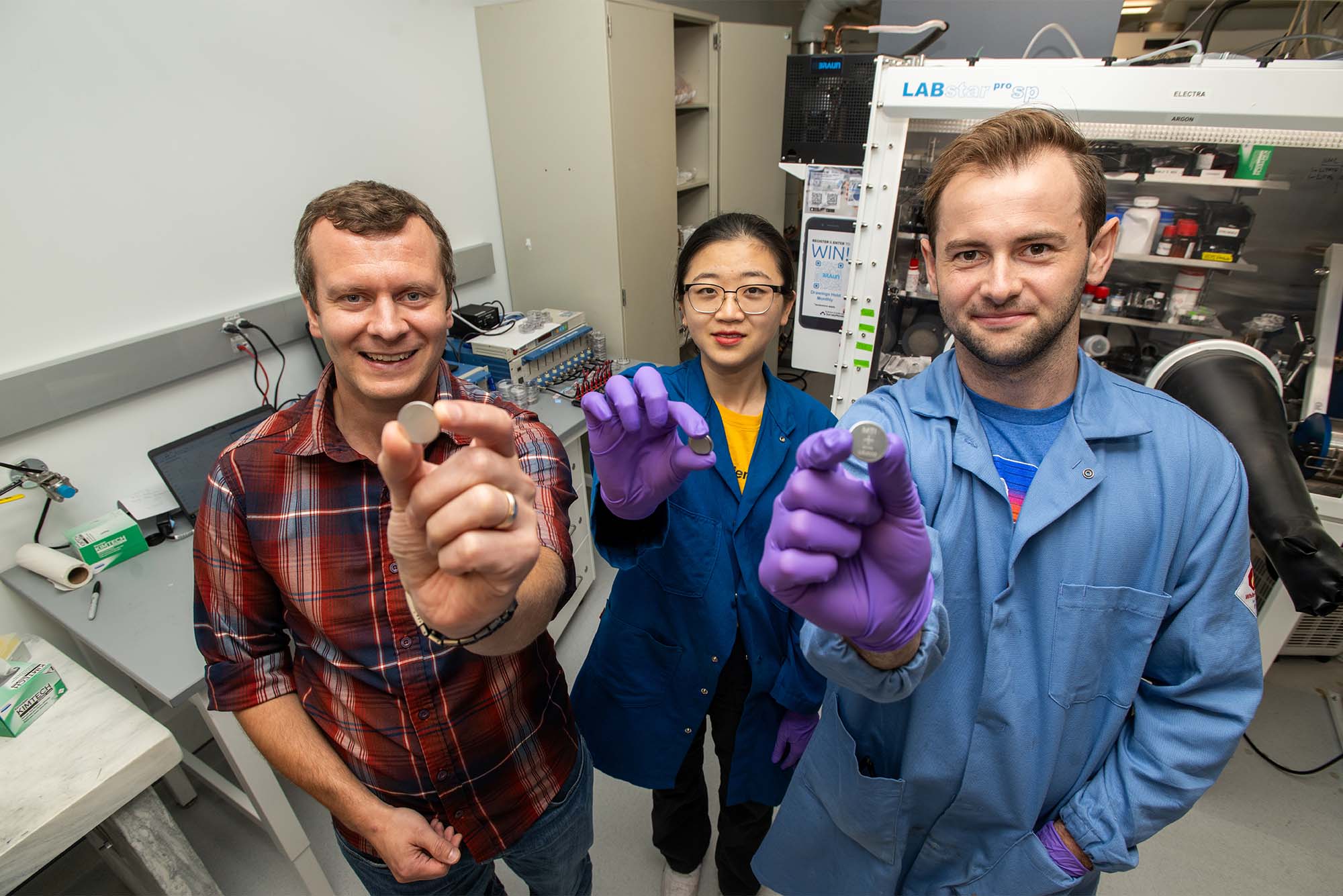 Photo: Three battery researchers hold up various coin shaped batteries in a scientific lab environment. They are all wearing lab coats