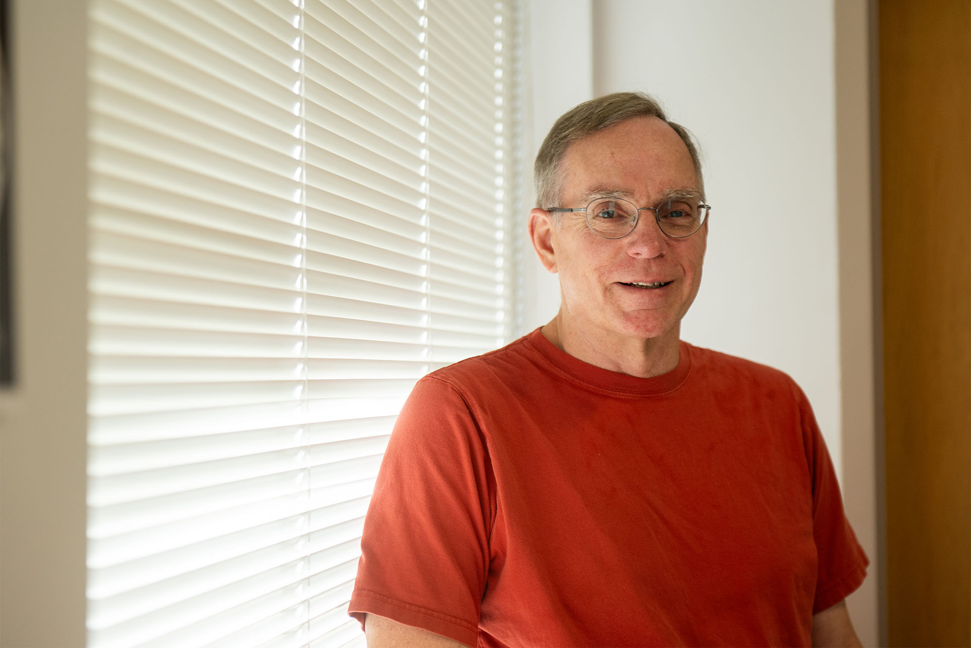 Photo: An older man wearing a red t shirt standing in front of a window with closed blinds