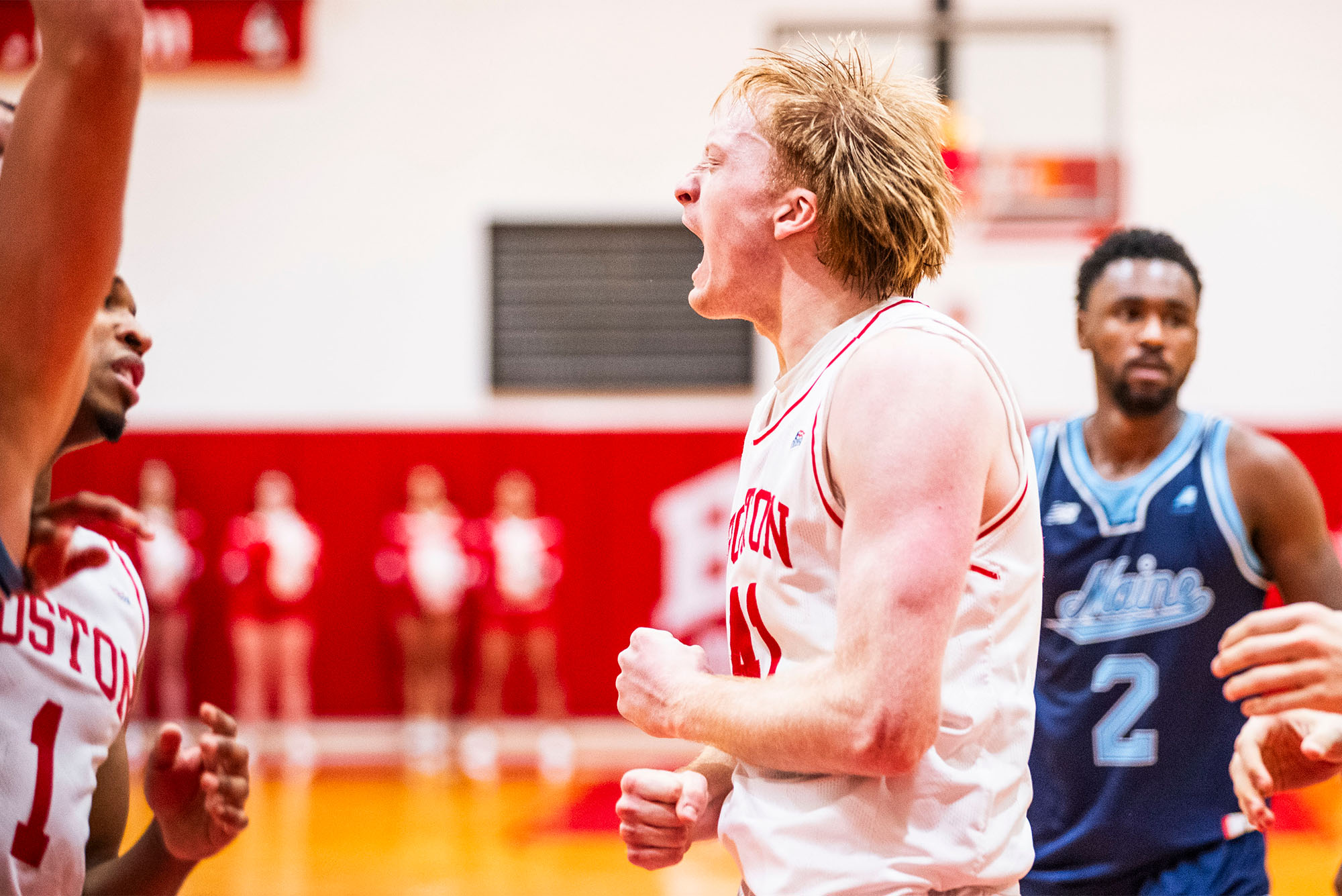 Photo: A college basketball player in a red and white jersey celebrates at a game. Other players are wearing dark blue jerseys. 